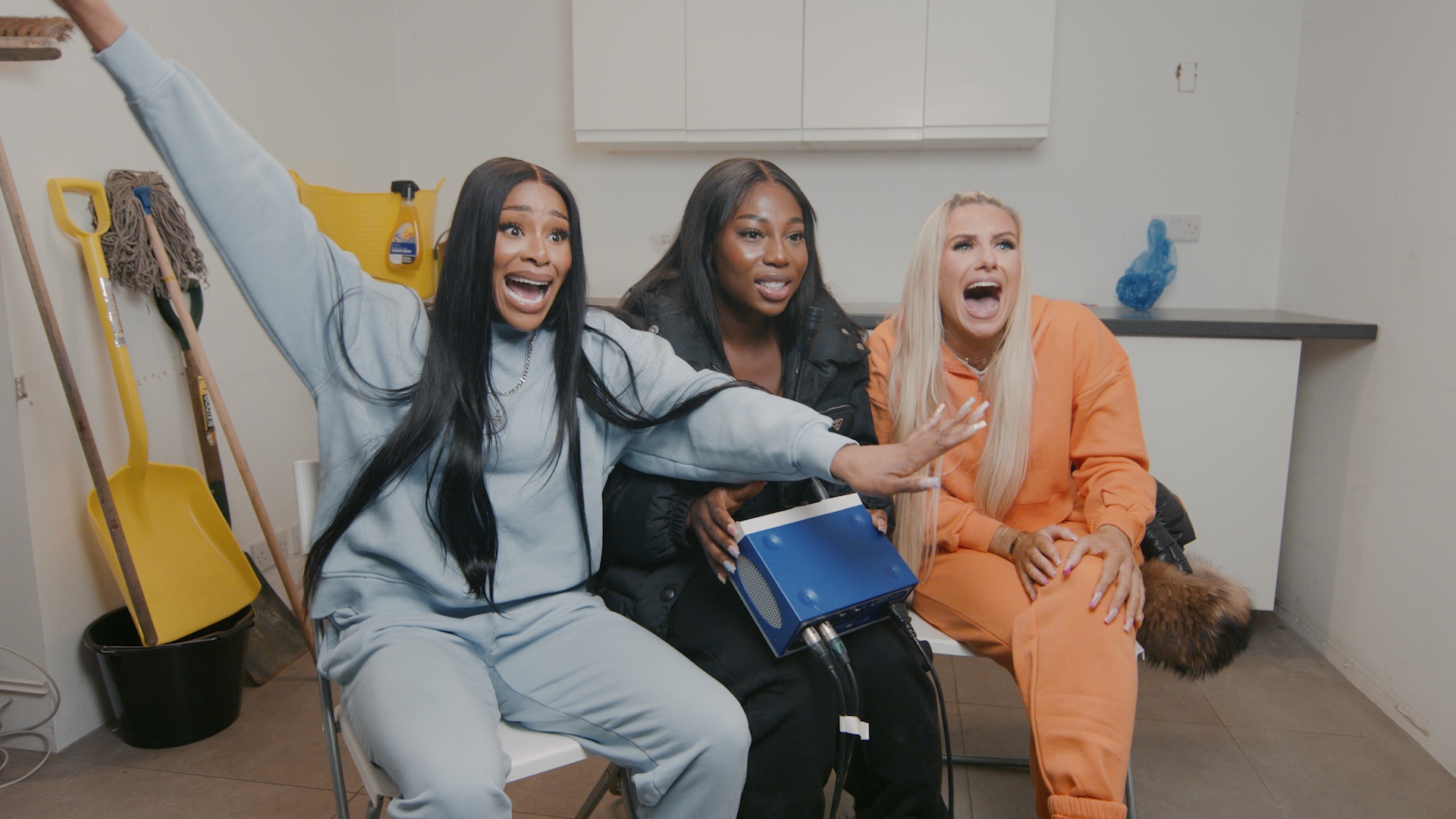 A scene from Channel 4's Tapped Out with three women sat on chairs in a utility room react to something they are seeing. 