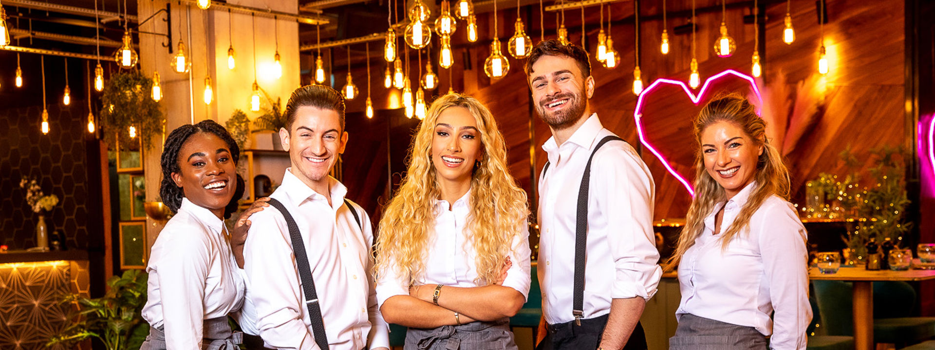 The five on-screen waiters from Teen First dates dressed in their waiting outfits of dark trousers, white shirts and grey aprons standing in front of a bar with a pink neon heart behind.